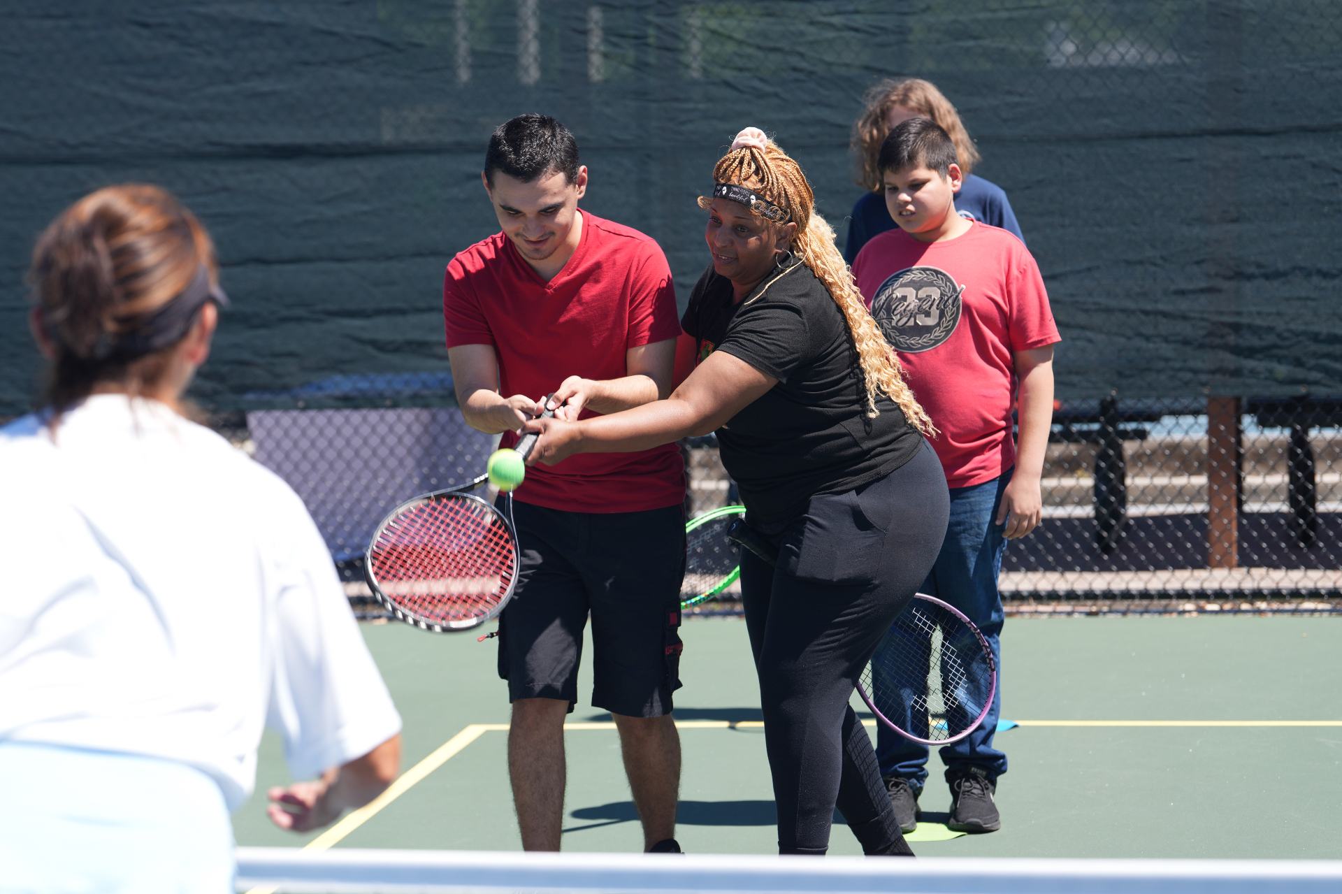 Coach helping a player swing through a tennis ball while teammates wait their turn